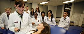 A White woman looks down at woman on examination table, as multiracial group of students look on.