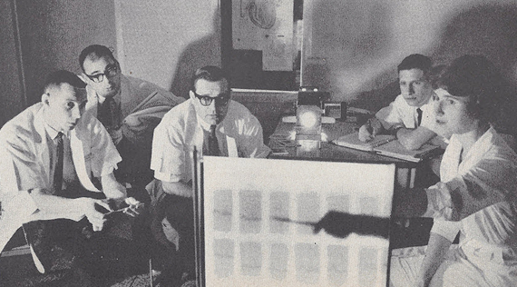 Four seated White men in lab coats look on as a White woman points towards a back lit board.